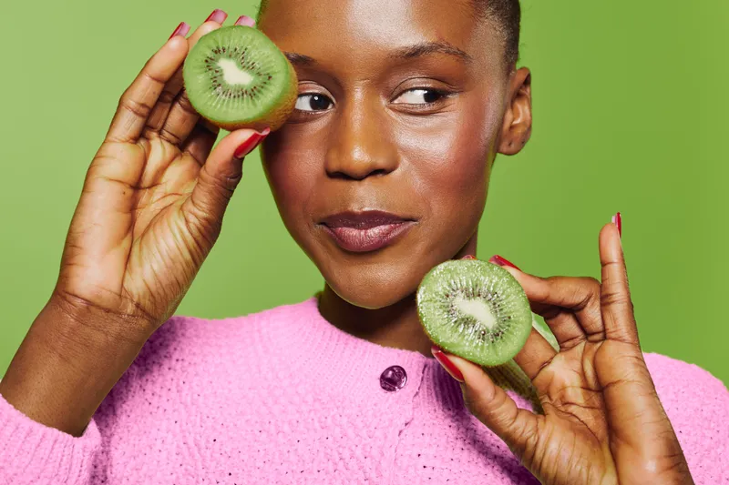 Young black woman with dark skin holds two halves of fresh kiwi near face, wearing pink sweater. Green background. Concept of healthy lifestyle, fruit, nutrition, and natural beauty.