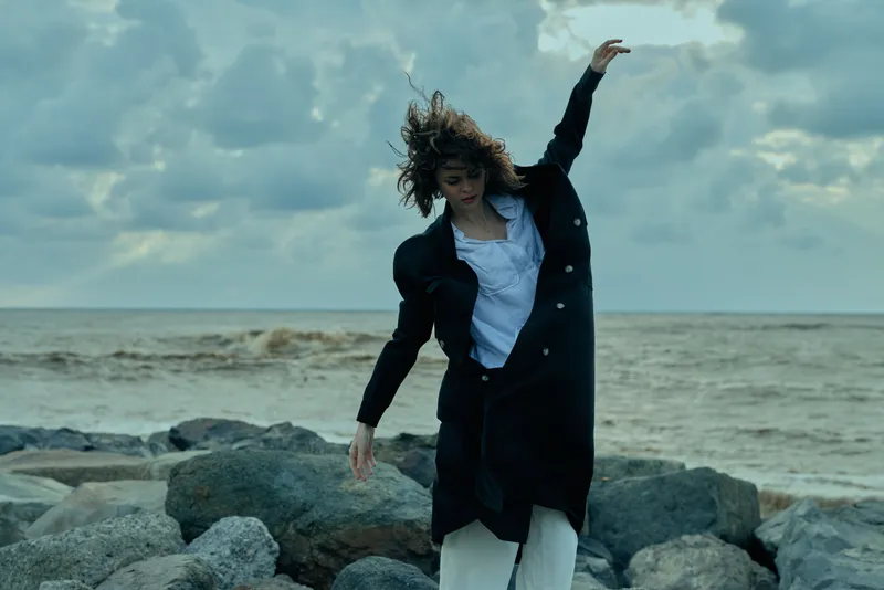 Fashion, motion, sea, wind, on rocky shoreline, a coat and white shirt move with the breeze as a figure strides by the water, delivering dynamic coastal style and a moody weather moment