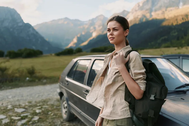 Woman with a backpack beside a car in a mountainous landscape, outdoors lifestyle scene. Casual pose, nature setting, sense of exploration, calm and hopeful mood.