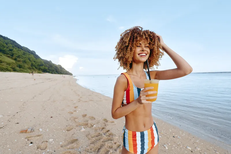 Smiling woman with curly hair on a sunny beach, holding a drink cup, wearing a striped swimsuit, sea waves behind her, relaxed summer vacation mood, natural light, outdoors