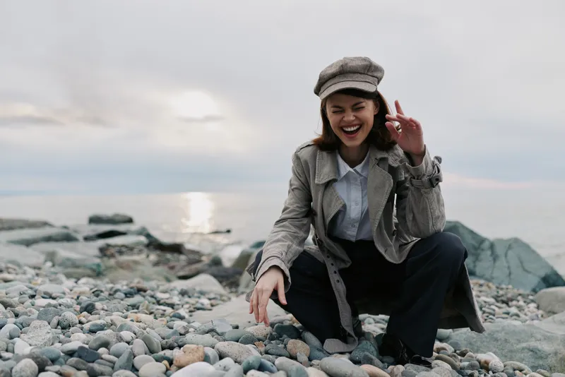 Smiling woman in stylish coat and hat crouches on rocky beach near water under cloudy sky. Casual autumn outfit and joyful mood captured outdoors by the sea during daytime.