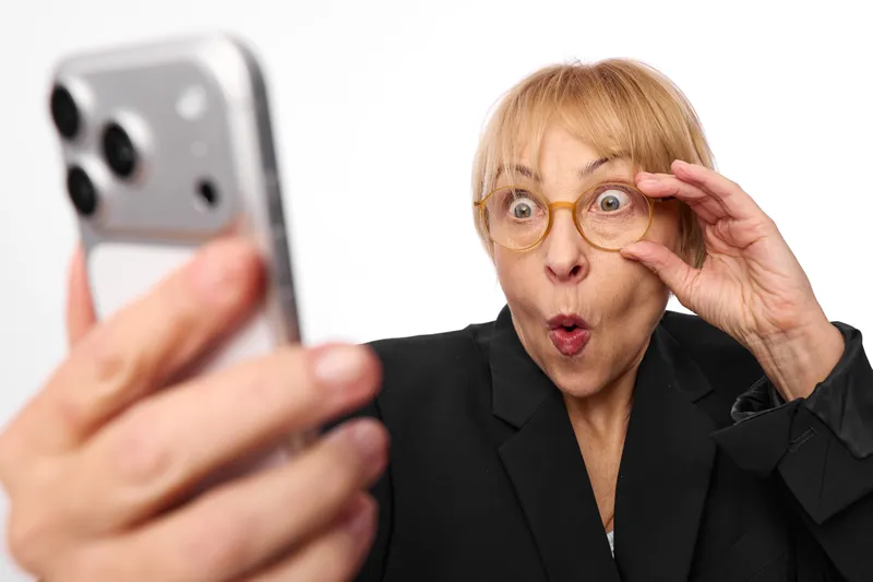 Surprised older woman in business attire holding a smartphone for a selfie, wearing round glasses, wide eyes and raised hand, close portrait, soft indoor light, copy space