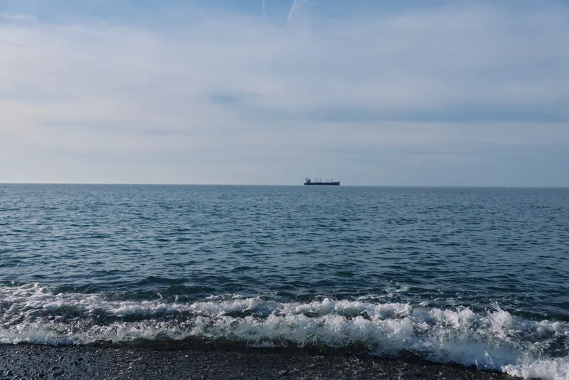 Sea and ocean scene with a distant ship on the horizon over calm water, gentle waves washing the shore, wide sky above, tranquil coastal view for travel and maritime exploration