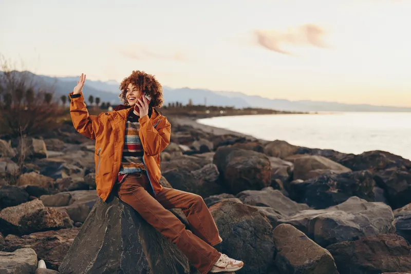 Woman lifestyle on beach at sunset, seated on rocks, relaxed pose with curly hair, orange jacket and jeans, travel leisure outdoors, soft warm light by sea, calm mood evening