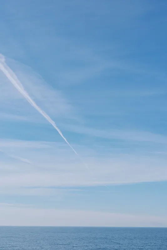 Sky and ocean meet at a calm horizon in clear blue light, with wispy clouds and a single contrail crossing the vast sky above tranquil water.