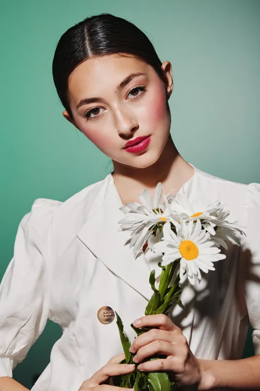 Young woman in white fashion outfit holds daisies at chest level against green background, showcasing flawless makeup and confident pose for a modern beauty, creative fashion concept.