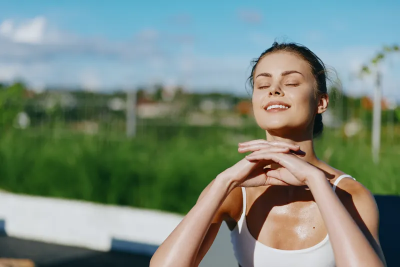 Cheerful woman outdoors, eyes closed, basking in sunshine, relaxed posture, warm breeze, casual athletic top, natural glow, sunlit moment, serene happiness, carefree lifestyle, vibrant colors,