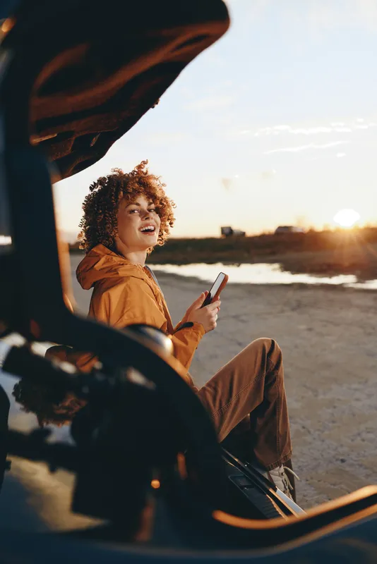 Woman lifestyle at beach sunset, curly hair, travel mood, relaxed laughing in an orange jacket by the coast, sandy shore and ocean, warm golden light, casual outdoors.