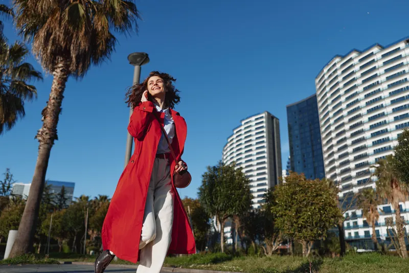 A stylish young woman in a bright red trench walks in a sunny city park, smiling as she carries a bag and enjoys an urban afternoon with palm trees and modern buildings.
