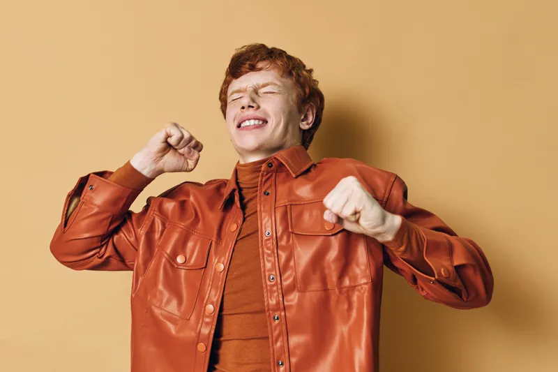 Young man with red hair wearing orange jacket stretching or yawning with closed eyes standing against beige background in casual clothing at studio.