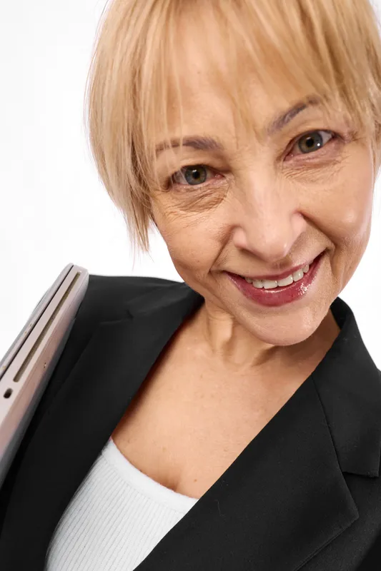 Senior woman portrait, smiling warmly, blonde short hair, studio white background, wearing blazer over white sweater, holding office folder, friendly expression, close up headshot