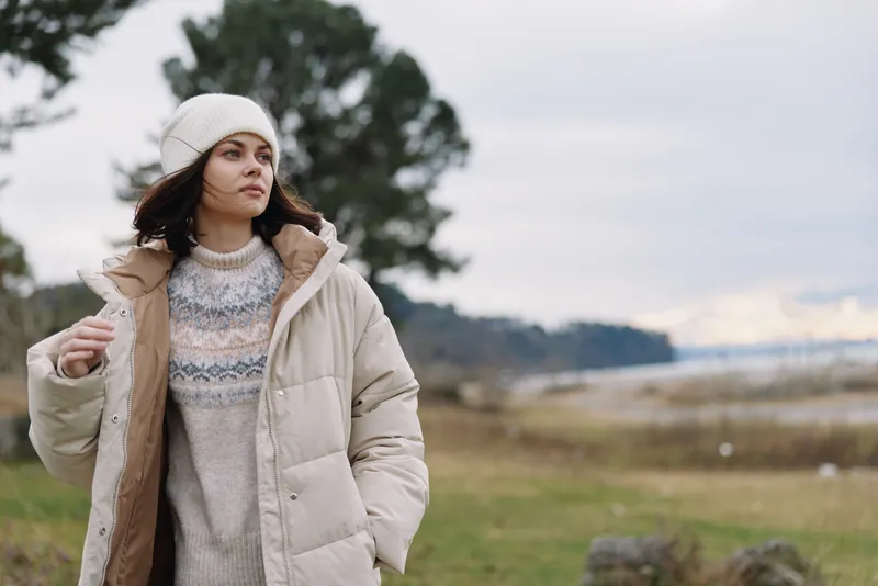 Woman in an oversized beige jacket and knit sweater stands outdoors in a windswept landscape, calm mood, soft light, moody sky, casual fashion moment