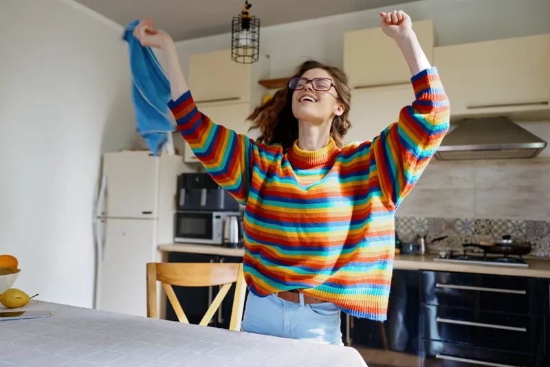 Bright, cheerful woman in a colorful striped sweater dances with laundry in a modern kitchen, arms raised, smiling. A candid lifestyle moment celebrating home energy and everyday joy.