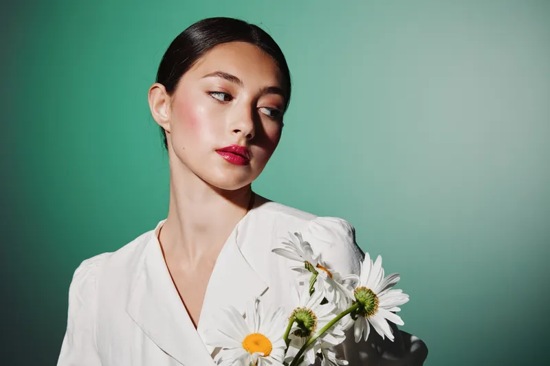 Young woman in elegant white outfit holding chamomile flowers while looking sideways. Studio fashion portrait with creative beauty concept and green studio background soft light.