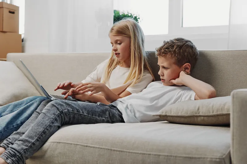 Two children sitting on sofa using laptop together in bright living room. Concept of childhood, learning, technology and sibling bonding at home.