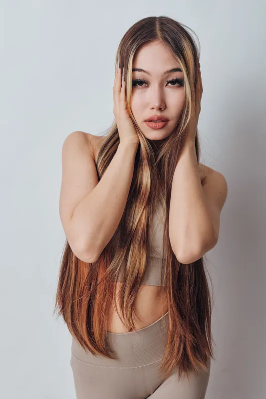 Young woman with long hair in beige top and pants posing with hands on face against plain background. Beauty, fashion, casual style and natural makeup for portrait photography.