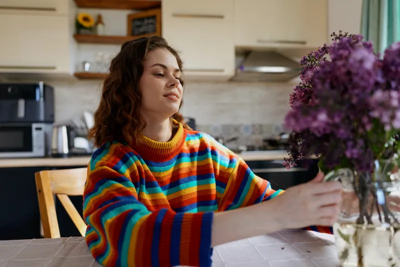 A woman sits at a sunny kitchen table, reaching toward a bouquet of flowers. A colorful striped sweater adds warmth to a bright, homey scene focused on lifestyle.