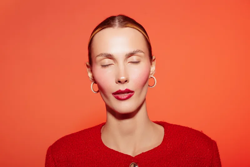 Close-up portrait of a woman with closed eyes wearing red lipstick and red sweater against orange background. Concept of beauty, makeup, fashion, and calm expression.