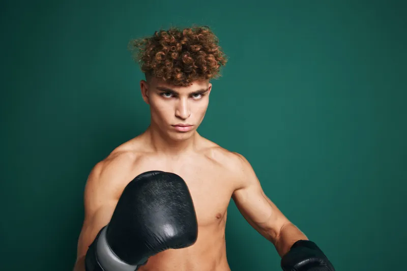 Young male boxer with curly hair wearing black gloves in fighting stance against green background for training and fitness concept.