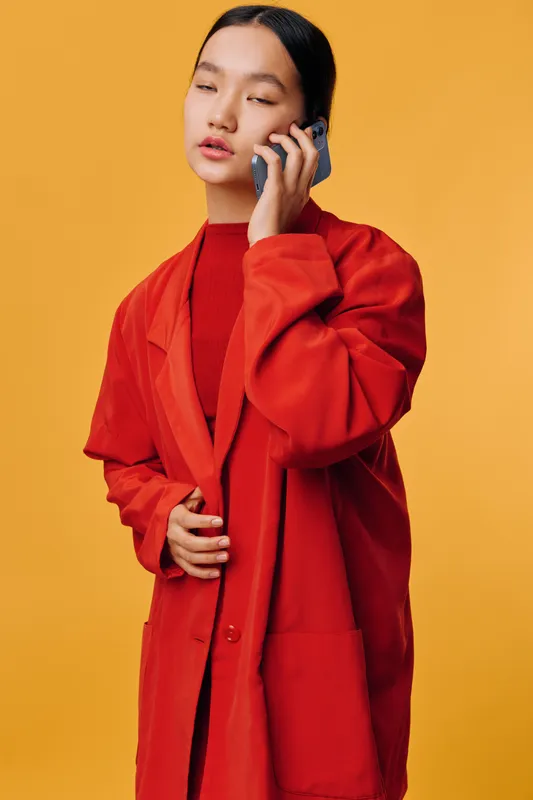 Young woman with long dark hair wearing red coat and sweater talking on phone with eyes closed on solid yellow background. Portrait, communication, fashion and lifestyle concept.