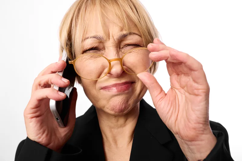 Elderly woman adjusting glasses while showing smile, holding phone near her ear, cheerful studio portrait on white background, relaxed hands and gaze, soft light