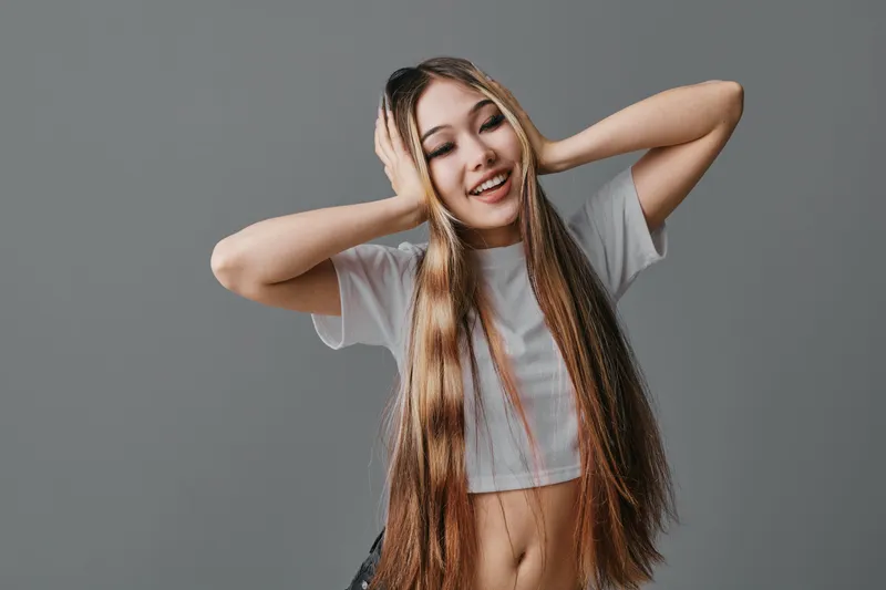 Young woman with long braided hair smiling and holding her head with hands on gray background. Casual clothing, happy emotion and hairstyle in studio closeup portrait.