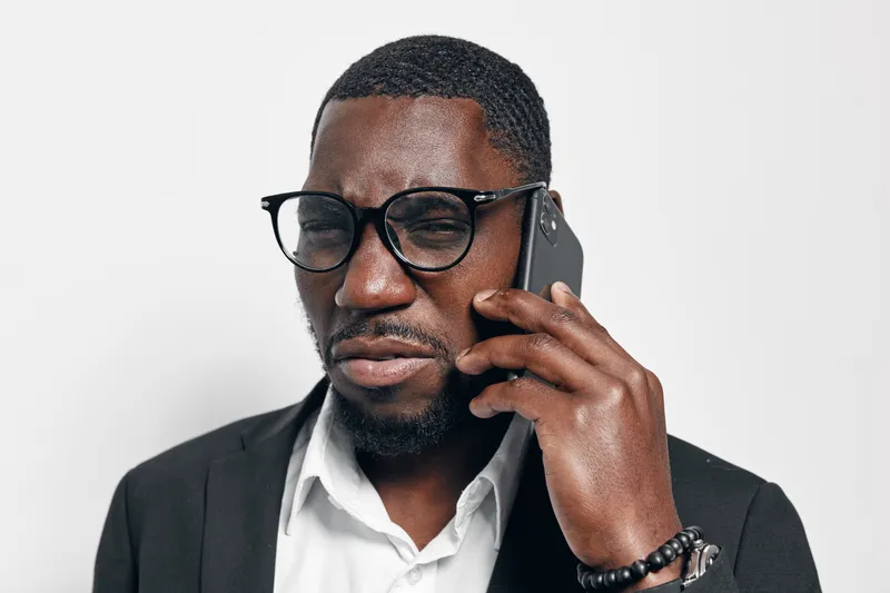 African American man with dark skin wearing glasses and suit talking on smartphone with serious expression isolated on white background in studio.