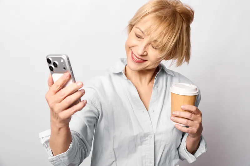 Blond woman smiling while holding smartphone and takeaway coffee cup, casual indoor portrait, using mobile communication, relaxed mood, copy space on blank background, morning lifestyle