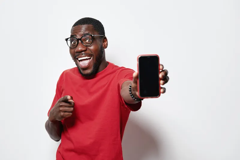 African American man with dark skin wearing glasses and red shirt smiling and pointing while showing smartphone with blank screen on white background.