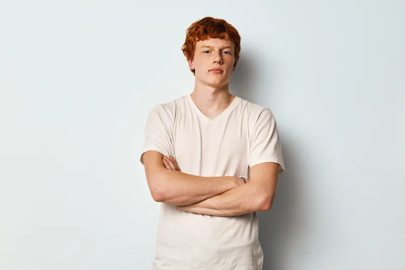 Young man with red hair and pale skin stands against white wall wearing plain white t shirt with arms crossed looking at camera in confident casual portrait studio shot.