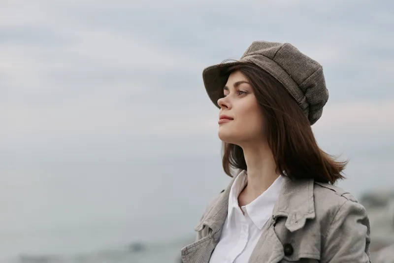 Young woman in a gray coat and checkered hat looks thoughtfully into the distance near the sea under a cloudy sky. Concept of contemplation, autumn fashion, and peaceful outdoor moments.