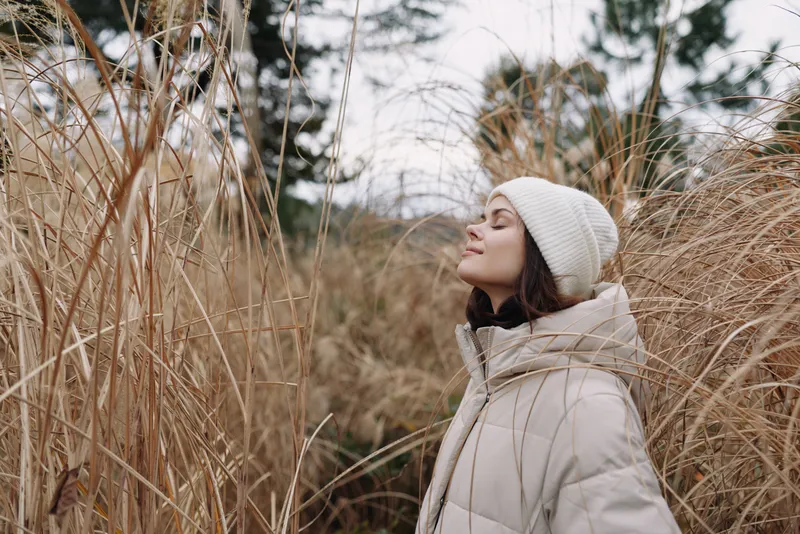 Woman in a warm coat and beanie stands among tall grasses outdoors, enjoying a serene winter moment in a natural landscape.