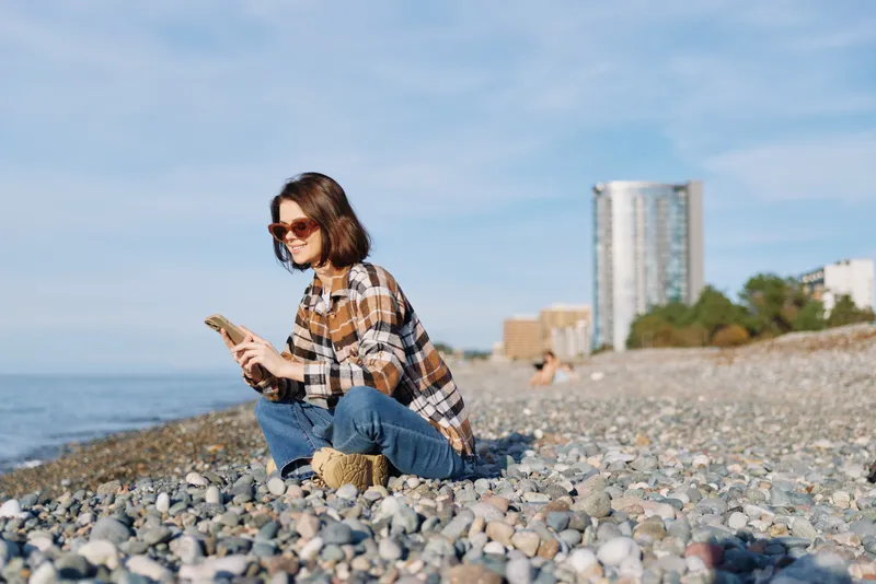Casual woman sitting on a pebble beach by the sea, checking her phone, wearing a plaid shirt and jeans, with distant city buildings and a bright sky, soft breeze and relaxed mood.