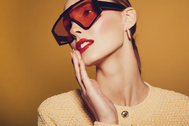 Stylish woman with oversized sunglasses and red lipstick poses with hand near face on yellow background. Fashion concept with modern accessories and confident expression.