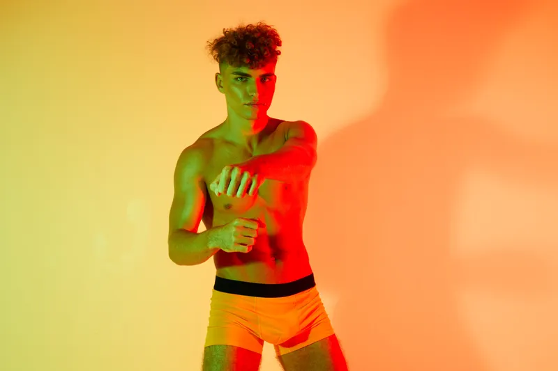 Young man with curly hair posing shirtless in colorful lighting, wearing orange shorts, showing confident expression and muscular body in studio with vibrant background.