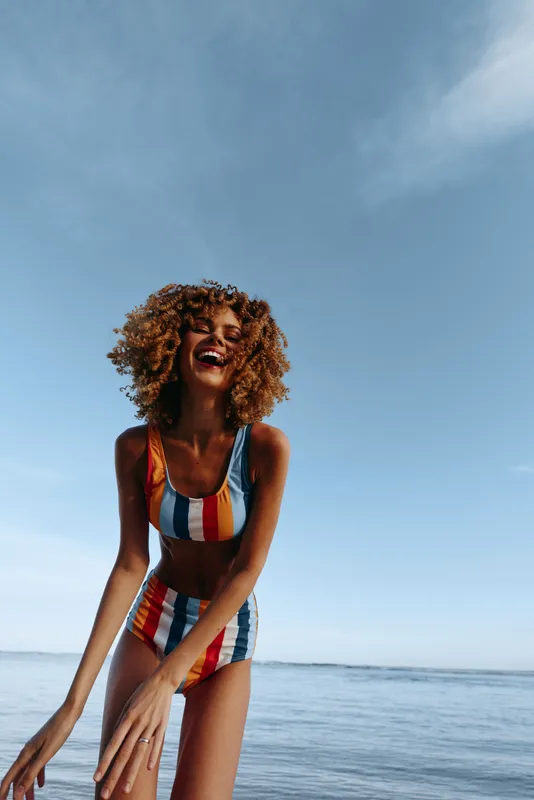 Smiling woman with curly hair in a striped swimsuit, standing by the seaside with ocean horizon behind, bright summer sunshine, soft breeze, relaxed joyful pose