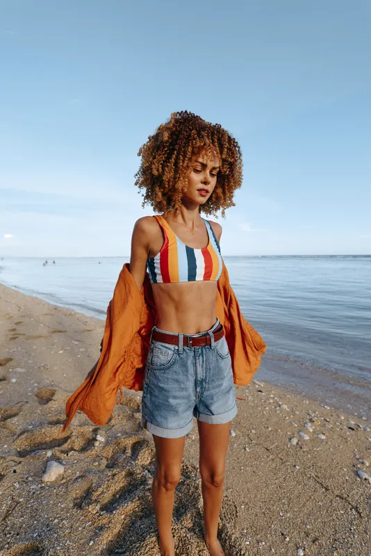 Woman beach ocean curly hair striped top denim jacket summer lifestyle, walking on shoreline with orange jacket, calm sea and blue sky, natural light, freedom mood outdoors today gently.
