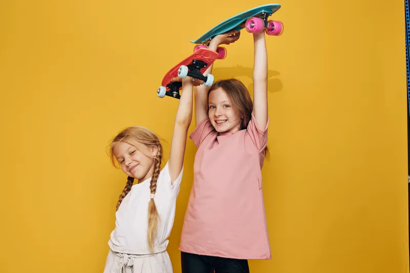 Two happy girls holding colorful skateboards above their heads smiling against yellow background. Concept for childhood, friendship, and leisure.