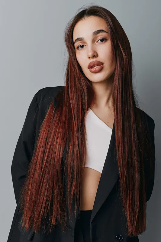 Woman with long red braids wearing black blazer and white top posing against gray background. Fashion portrait with natural makeup and confident expression for style and beauty.