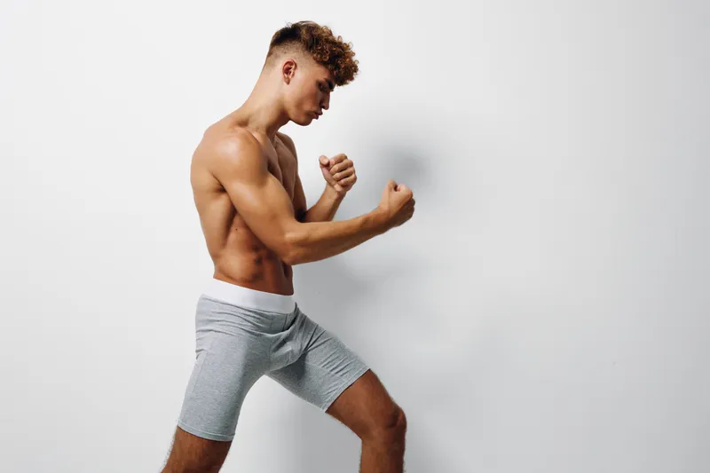 Young muscular man in gray shorts posing in fighting stance with fists clenched, showing strength and fitness against white background in studio. Athletic body and focused expression.
