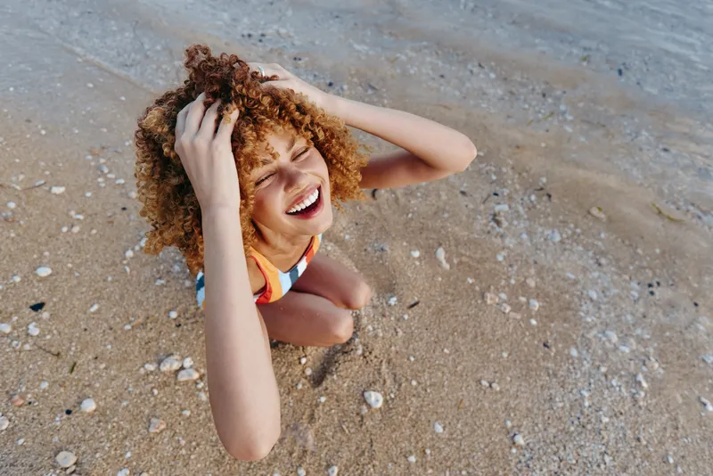 Young curly-haired woman in a colorful swimsuit laughs joyfully while sitting on a sandy beach near the water's edge on a bright day.