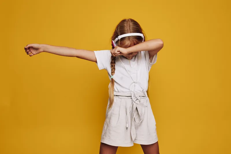Girl in headphones wearing white outfit performs dab dance move on yellow background. Concept of childhood, music, and playful expression.