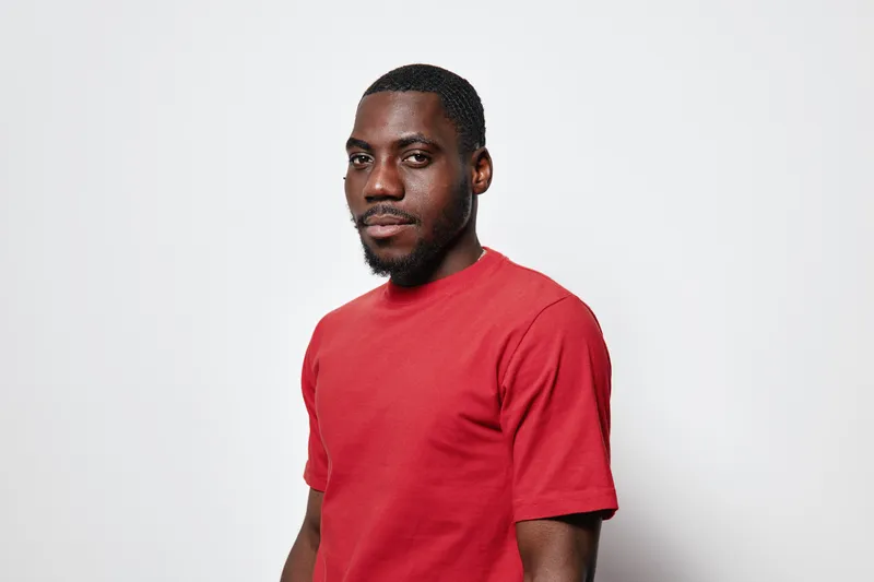 African American man with dark skin wearing red t shirt standing against white background looking at camera with confident expression and relaxed posture in studio.