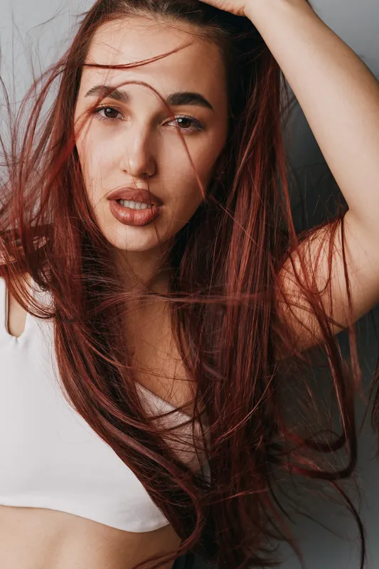 Woman with long red hair and natural makeup posing in white crop top against gray background. Closeup portrait with loose hair strands and confident facial expression in studio lighting.