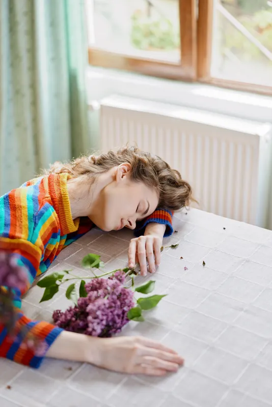 Woman resting at home, lying on a table with a bouquet of flowers, wearing a colorful striped sweater, cozy and calm mood, sunlight through a window, intimate lifestyle scene.