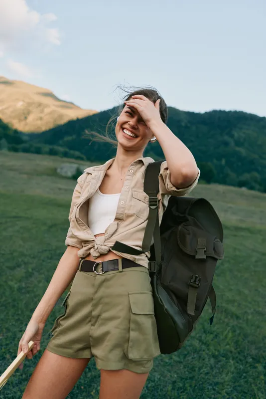 Woman traveler with backpack on a grassy hillside, enjoying outdoor life, casual attire, bright day, and a relaxed confident mood, capturing everyday adventure in nature