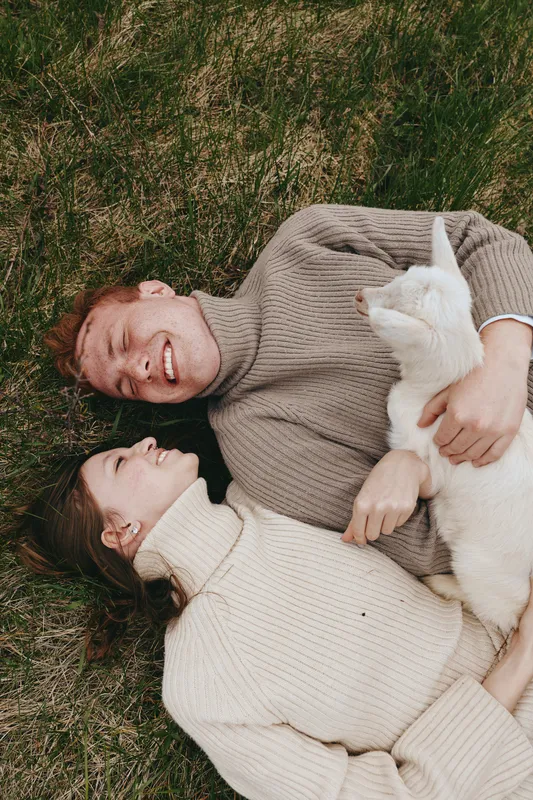Happy couple lying on grass in cozy sweaters, sharing laughter with a white cat, a heartfelt lifestyle moment outdoors, relaxed affectionate atmosphere, candid and warm.