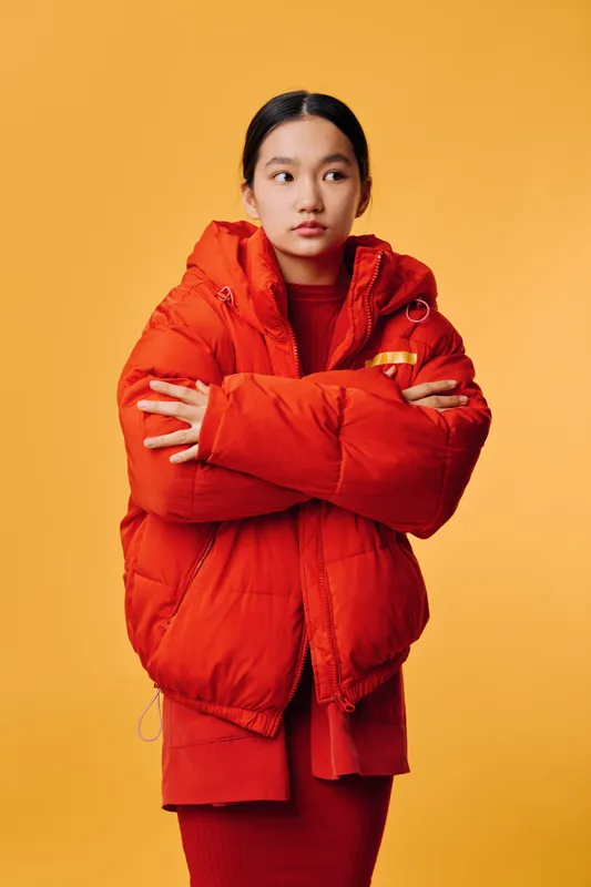 Young woman in red puffer jacket and sweater standing with crossed arms against orange background. Casual fashion, winter clothing, serious expression and Asian female portrait with studio lighting.