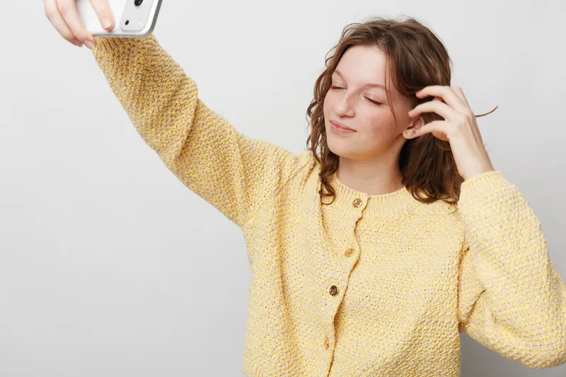 Young woman in yellow knitted sweater takes selfie with smartphone, smiling gently with closed eyes. Casual lifestyle, social media, and self-expression concept on plain background.