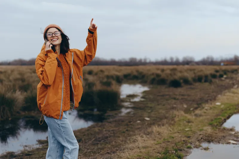 Woman lifestyle outdoors in nature, wearing orange jacket and jeans, standing on a muddy path by a river, smiling with raised hands, casual travel mood in overcast daylight.